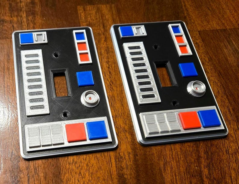Op de afbeelding: Twee zwarte lichtschakelaars met een zilveren en grijs ontwerp. De afdekkingen hebben verschillende knoppen en schakelaars, waaronder rode en blauwe knoppen. De afdekkingen zijn ontworpen om eruit te zien als een bedieningspaneel van een ruimteschip.