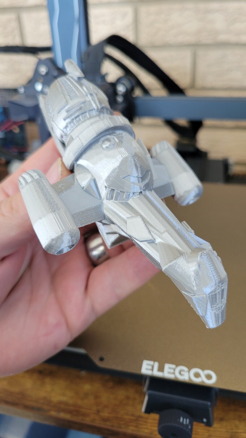 May include: A silver-colored, 3D-printed model of a spaceship, held in a hand. The model has a detailed design with wings and a central body. The image is taken indoors, with a 3D printer visible in the background.