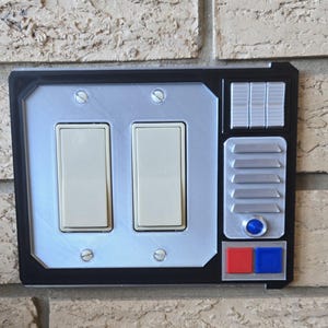 May include: A white light switch plate with two toggle switches and a black border. The plate has a silver panel with a blue button and red and blue buttons below it.