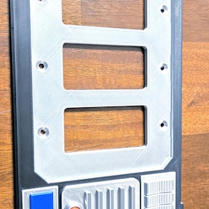 May include: A rectangular, 3D-printed panel with three rectangular cutouts. The panel has a silver-colored central section and a black border. It features a series of small, detailed sections with orange, blue, and white accents, and an orange circular detail.