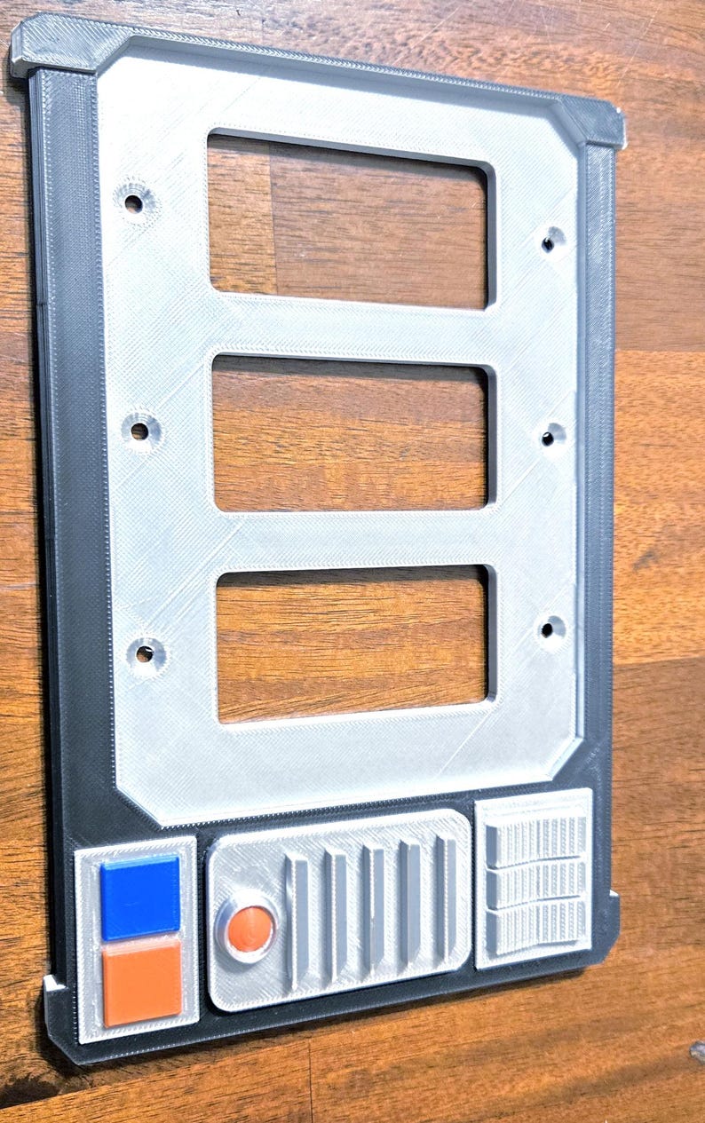 May include: A rectangular, 3D-printed panel with three rectangular cutouts. The panel has a silver-colored central section and a black border. It features a series of small, detailed sections with orange, blue, and white accents, and an orange circular detail.