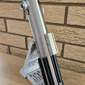 May include: A silver and black lightsaber replica, displayed on a gray stand. The lightsaber features detailed metallic components, including a control panel and emitter. The stand is designed to hold the lightsaber at an angle.