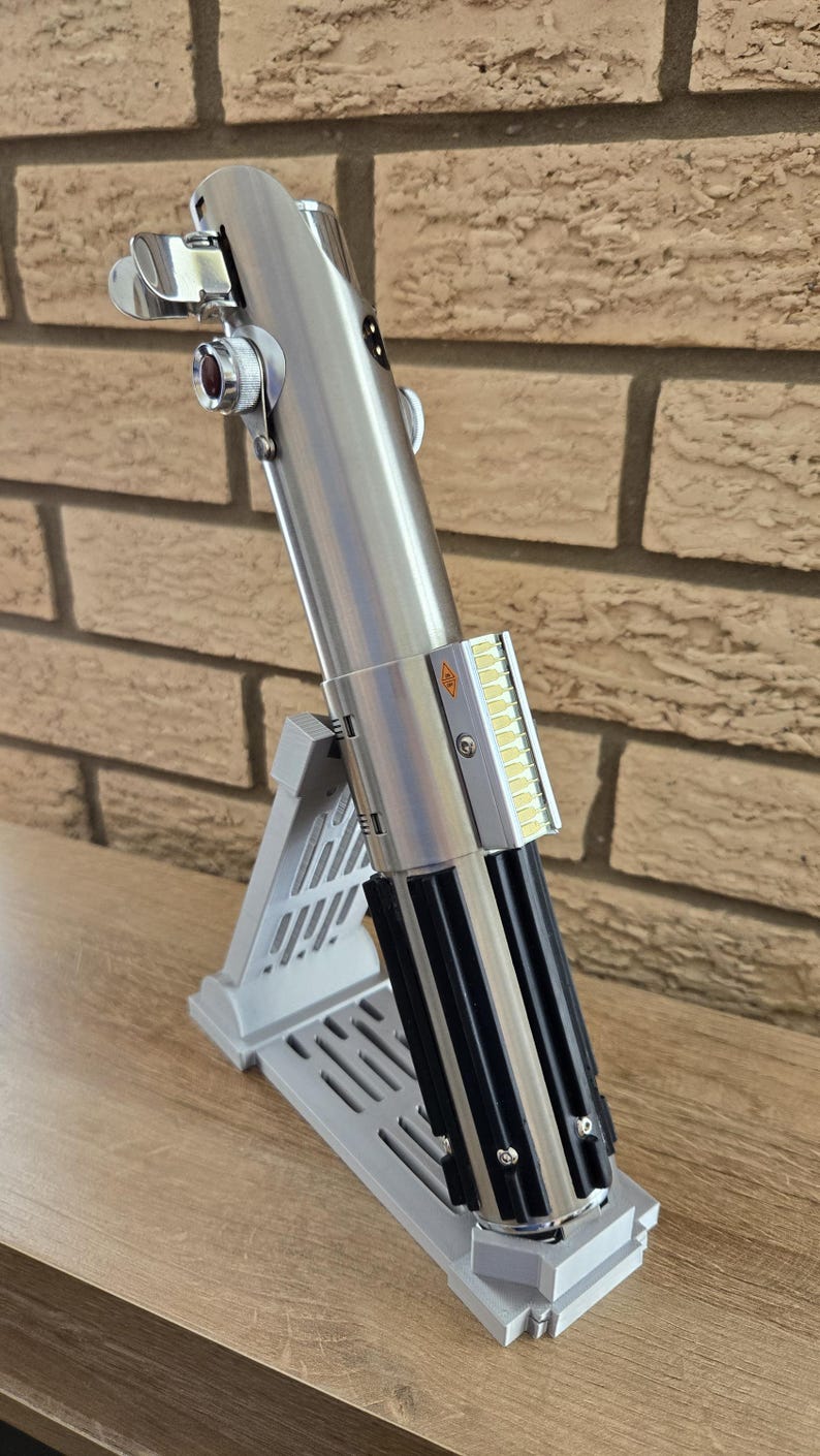 May include: A silver and black lightsaber replica, displayed on a gray stand. The lightsaber features detailed metallic components, including a control panel and emitter. The stand is designed to hold the lightsaber at an angle.