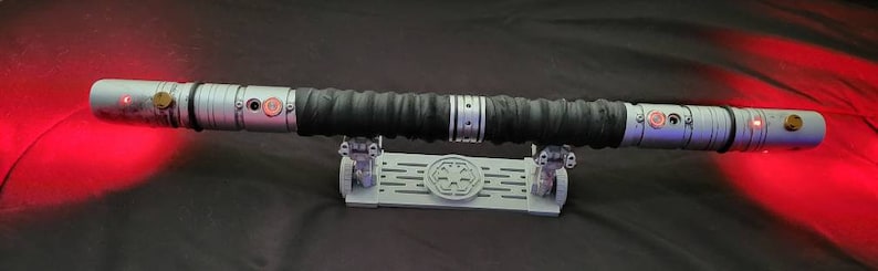 May include: A silver lightsaber with a black handle and red lightsabers on each end. The lightsaber is on a gray stand with a symbol on it.