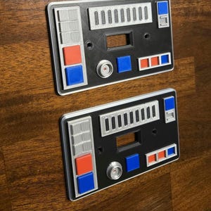 Op de afbeelding: Twee zwarte en zilveren lichtschakelaars met rode, blauwe en witte knoppen. De covers hebben een Star Wars-ge&iuml;nspireerd ontwerp.