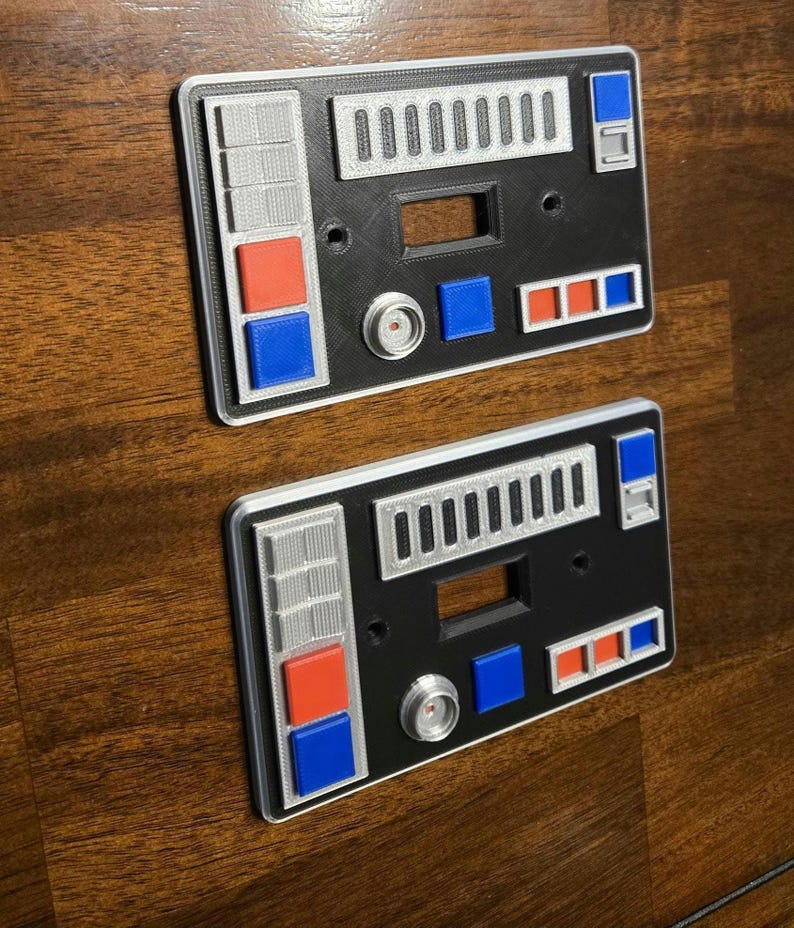 Op de afbeelding: Twee zwarte en zilveren lichtschakelaars met rode, blauwe en witte knoppen. De covers hebben een Star Wars-ge&iuml;nspireerd ontwerp.