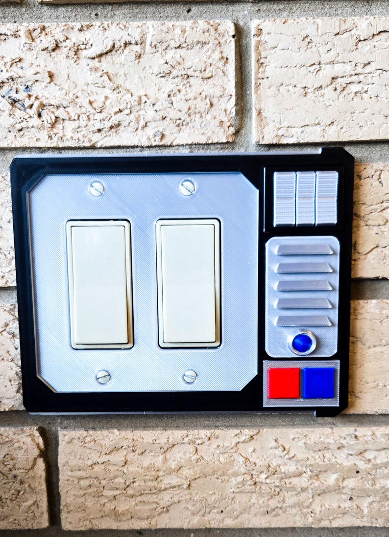 May include: A black light switch cover with a silver and blue control panel. The control panel has a red and blue button, a round blue button, and five silver rectangular buttons.