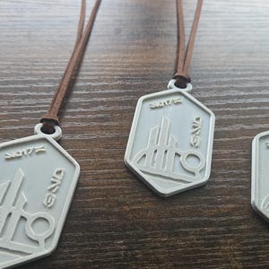 Puede incluir: Collares con colgantes plateados y cordones marrones. Cada colgante tiene una forma geom&eacute;trica con un dise&ntilde;o estilizado y texto. Los collares se exhiben sobre una superficie de madera.