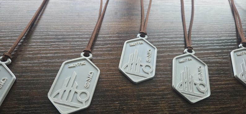 Puede incluir: Collares con colgantes plateados y cordones marrones. Cada colgante tiene una forma geom&eacute;trica con un dise&ntilde;o estilizado y texto. Los collares se exhiben sobre una superficie de madera.