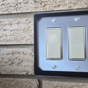 May include: A black light switch cover with a silver panel featuring two toggle switches. The cover has a futuristic design with a silver panel on the right side with a blue button and red and blue buttons below it.