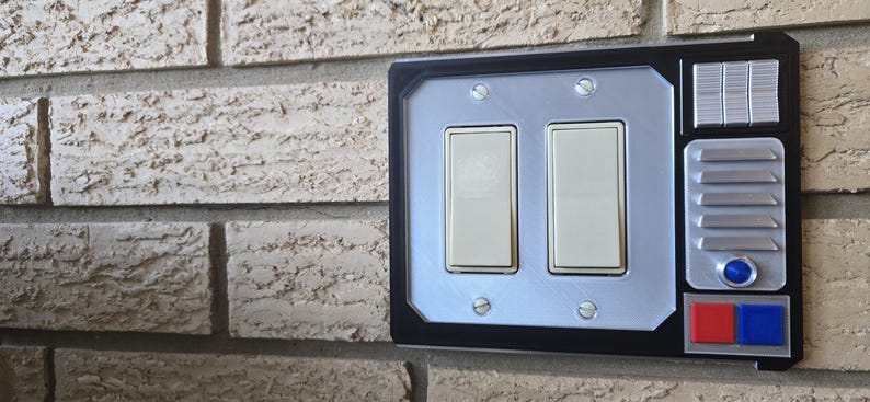 May include: A black light switch cover with a silver panel featuring two toggle switches. The cover has a futuristic design with a silver panel on the right side with a blue button and red and blue buttons below it.