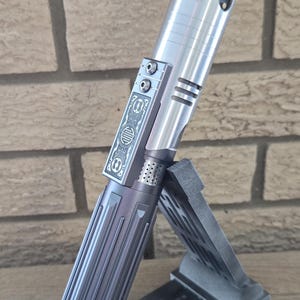 Op de afbeelding: Een zilveren lightsaber-handvat met een zwarte grip en een decoratief ontwerp aan de zijkant. Het handvat staat op een zwarte standaard.