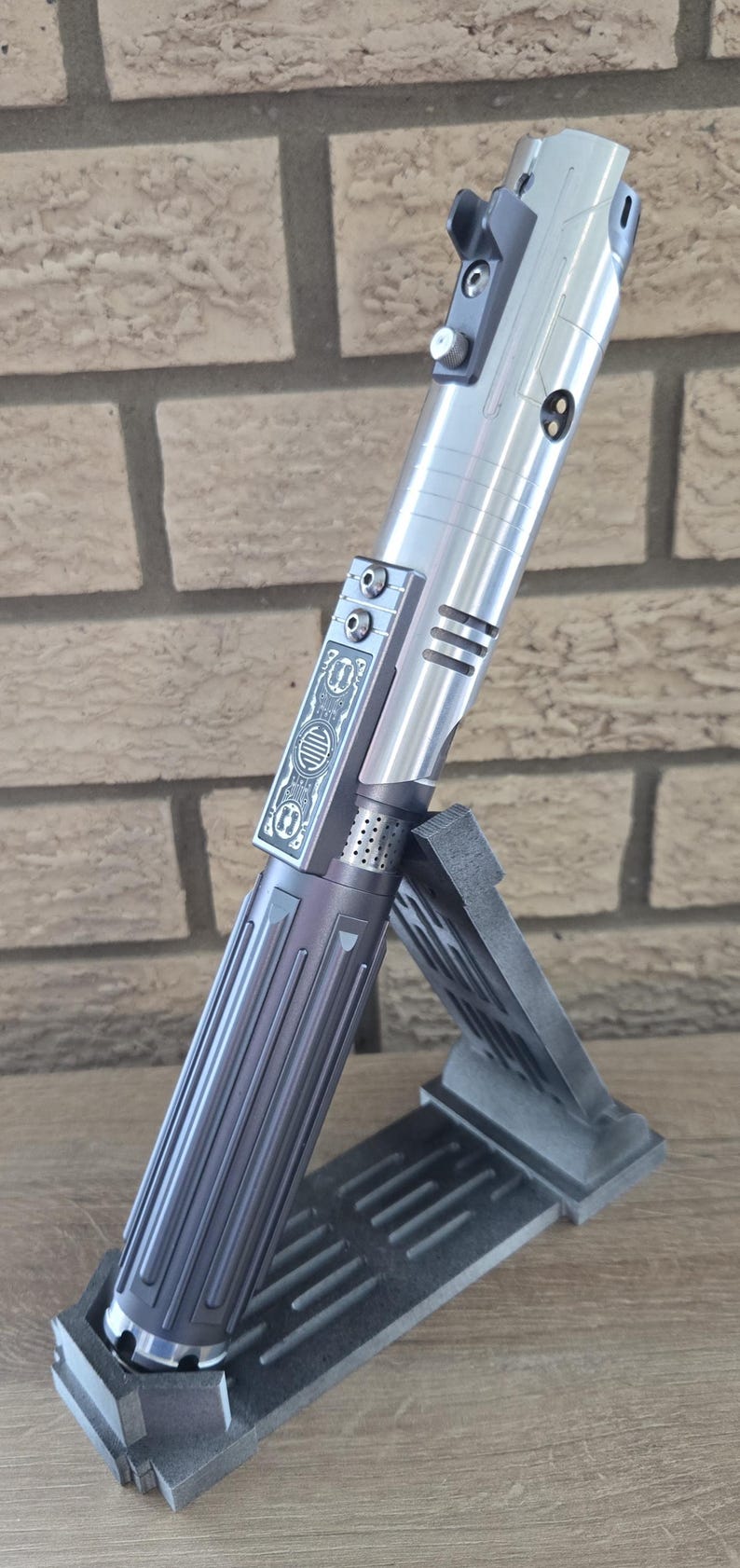 Op de afbeelding: Een zilveren lightsaber-handvat met een zwarte grip en een decoratief ontwerp aan de zijkant. Het handvat staat op een zwarte standaard.
