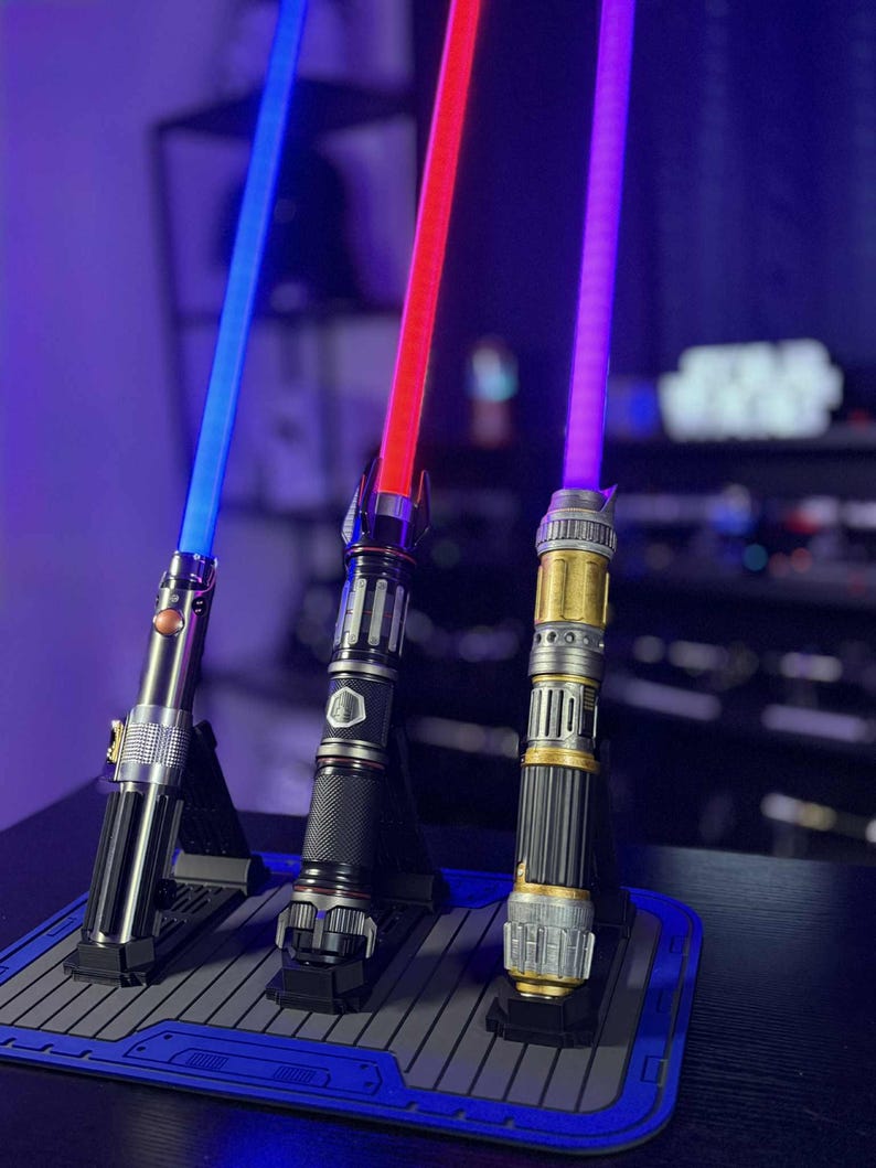 May include: Three illuminated lightsabers with blue, red, and purple blades are displayed on a dark surface. The hilts are silver, black, and gold, each with unique designs. The lightsabers are standing on individual black stands.