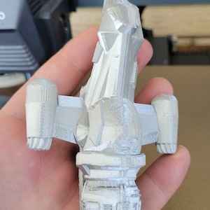 May include: A light gray 3D-printed model of a spaceship, held in a hand. The model has detailed features, including wings, a cockpit, and engine components. The object is made of a light gray plastic material.