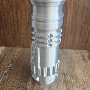 May include: A silver-colored lighter with a blue and red top, resembling a lightsaber. The lighter has a detailed design with ridges and grooves.