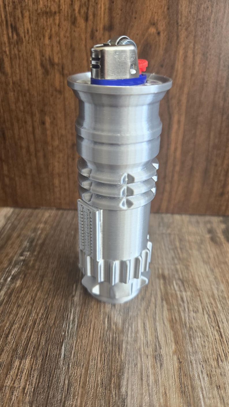 May include: A silver-colored lighter with a blue and red top, resembling a lightsaber. The lighter has a detailed design with ridges and grooves.