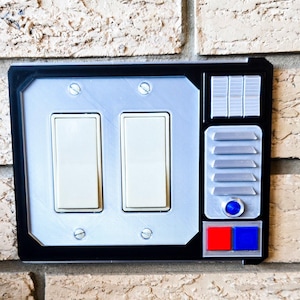 May include: A black and silver light switch cover with a futuristic design. It features two toggle switches, a series of buttons, and a red and blue button.