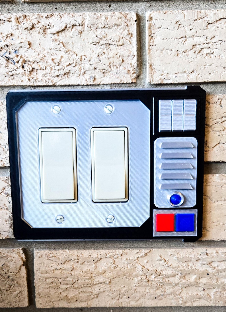 May include: A black and silver light switch cover with a futuristic design. It features two toggle switches, a series of buttons, and a red and blue button.