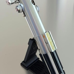 May include: A silver lightsaber replica with black accents, displayed on a black stand. The lightsaber features detailed metallic components and a textured grip. The stand is angled, showcasing the prop's design.