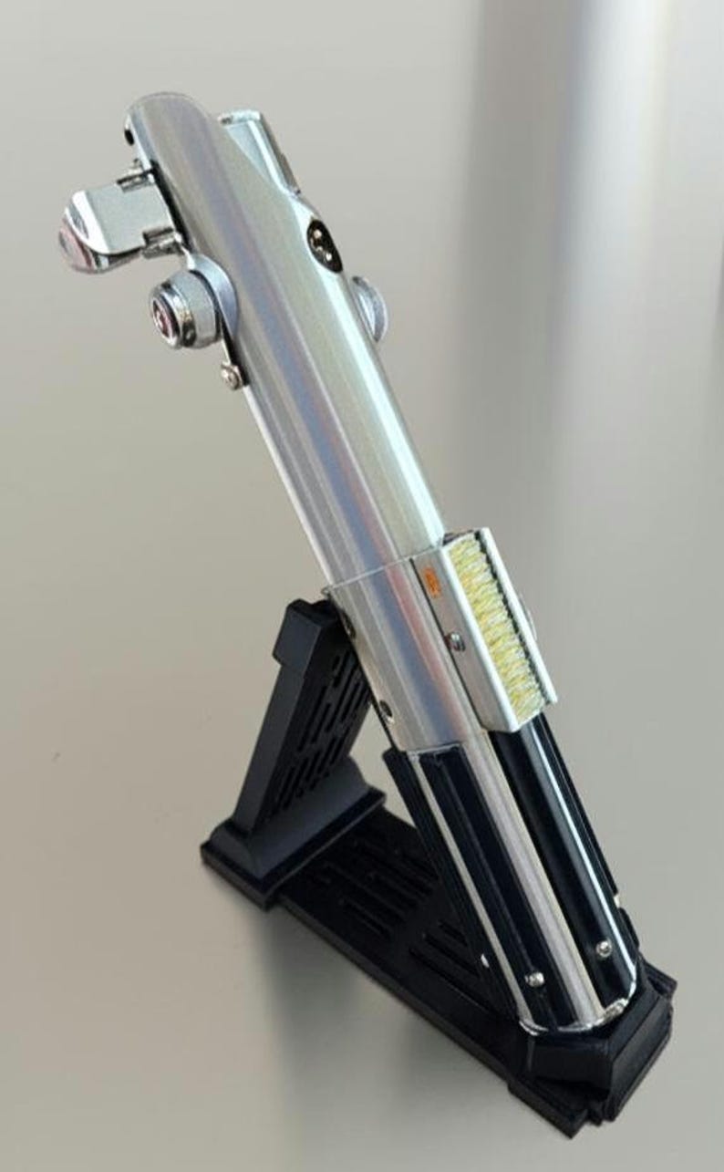 May include: A silver lightsaber replica with black accents, displayed on a black stand. The lightsaber features detailed metallic components and a textured grip. The stand is angled, showcasing the prop's design.