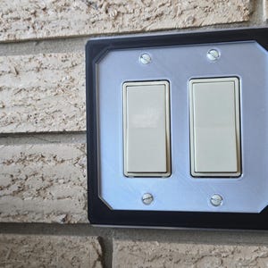 May include: A black and silver light switch plate with two toggle switches, a button, and a panel with a series of vertical lines. The plate has red and blue accents.