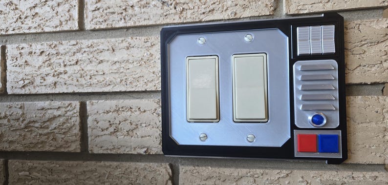 May include: A black and silver light switch plate with two toggle switches, a button, and a panel with a series of vertical lines. The plate has red and blue accents.