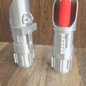 May include: Two silver lighters shaped like lightsabers. One lighter is open and shows a red lighter inside. The lighters are on a wood surface.