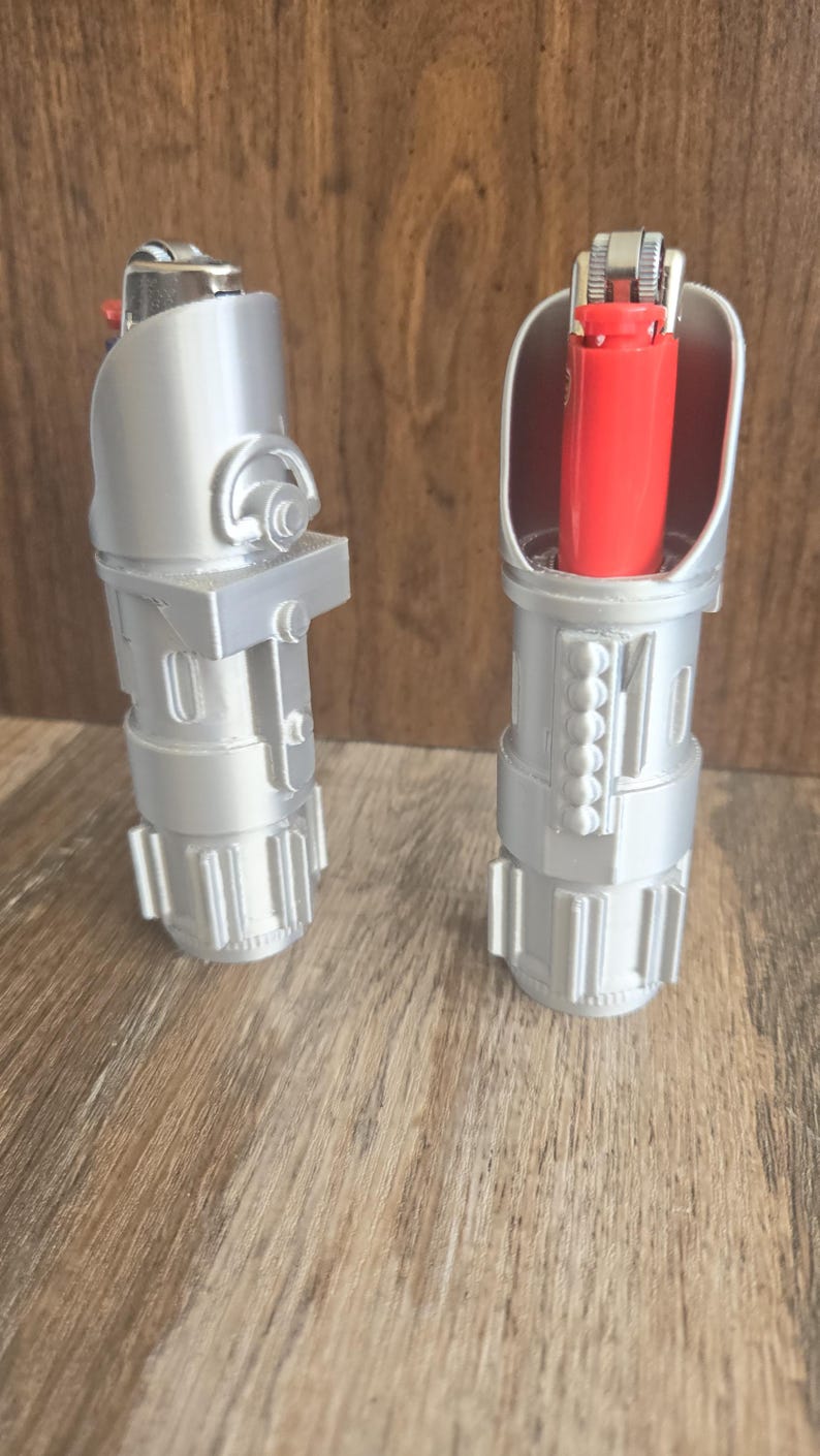 May include: Two silver lighters shaped like lightsabers. One lighter is open and shows a red lighter inside. The lighters are on a wood surface.