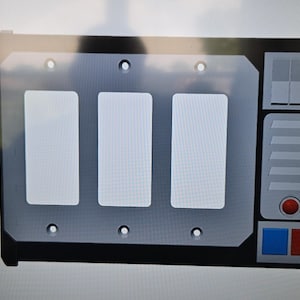 May include: A black and gray control panel with three white rectangular buttons. The panel has a silver faceplate with a series of small holes and a section with a red button and blue and red squares.