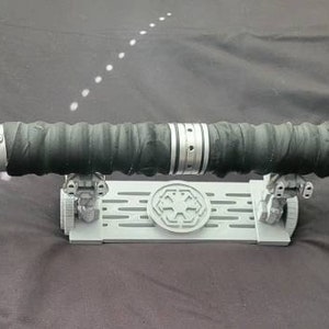 May include: A black lightsaber with a silver handle and red lights on each end. The lightsaber is on a white stand with a black symbol.