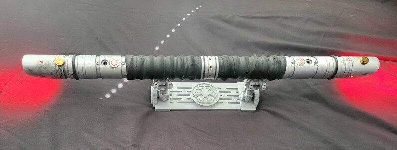 May include: A black lightsaber with a silver handle and red lights on each end. The lightsaber is on a white stand with a black symbol.