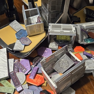 May include: A collection of small, gray, metal boxes with intricate designs. The boxes are open and contain various small, colorful tokens. The tokens are scattered around the boxes on a wooden surface.