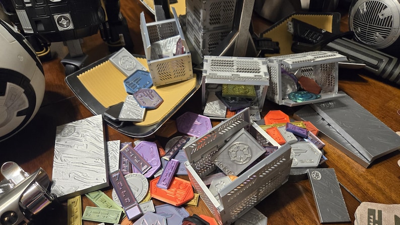 May include: A collection of small, gray, metal boxes with intricate designs. The boxes are open and contain various small, colorful tokens. The tokens are scattered around the boxes on a wooden surface.