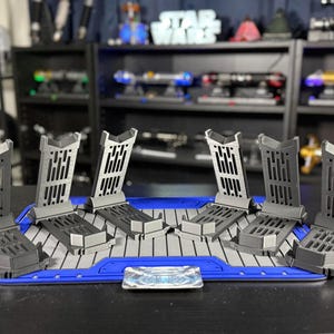 May include: A set of six gray lightsaber display stands arranged on a blue and gray platform. The stands have a unique design with vertical slots and are designed to hold lightsabers. A small, rectangular emblem is centered on the platform.