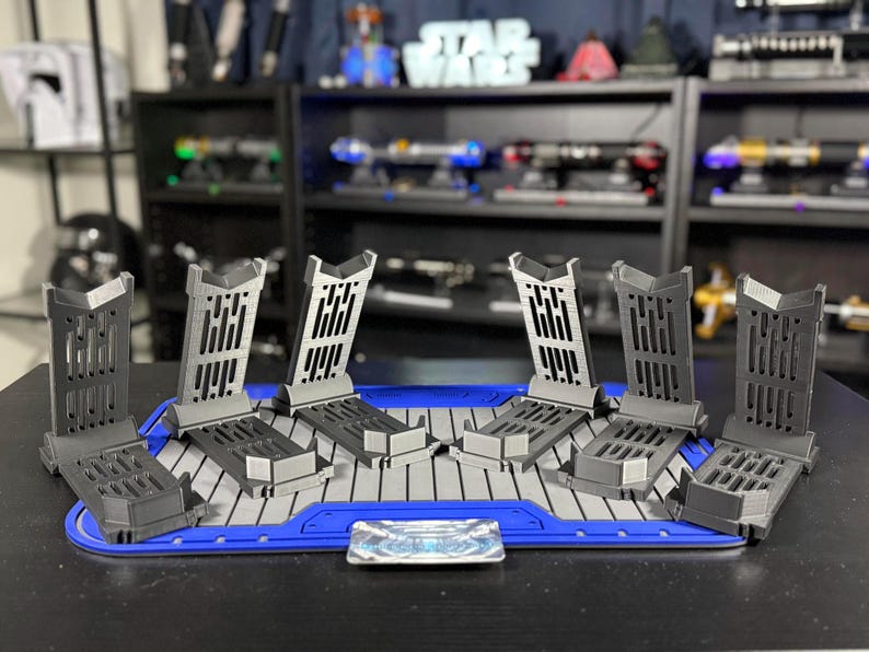 May include: A set of six gray lightsaber display stands arranged on a blue and gray platform. The stands have a unique design with vertical slots and are designed to hold lightsabers. A small, rectangular emblem is centered on the platform.