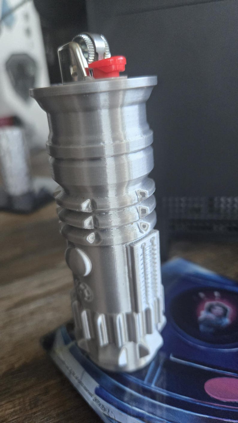 May include: A silver lightsaber lighter with a red button on top. The lighter is on a blue and black background.