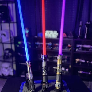 K&ouml;nnte beinhalten: Drei Lichtschwerter mit leuchtenden Klingen in Blau, Rot und Lila. Die Griffe sind silber, schwarz und goldfarben, jedes mit einzigartigen Designs. Sie werden auf einer blau-grauen Matte mit dunklem Hintergrund pr&auml;sentiert.