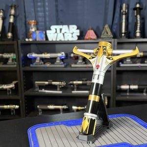 May include: A gold, silver, and black lightsaber is displayed on a stand. The lightsaber has a crossguard design and is surrounded by other lightsabers on shelves. The Star Wars logo is visible in the background.