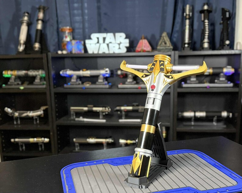May include: A gold, silver, and black lightsaber is displayed on a stand. The lightsaber has a crossguard design and is surrounded by other lightsabers on shelves. The Star Wars logo is visible in the background.