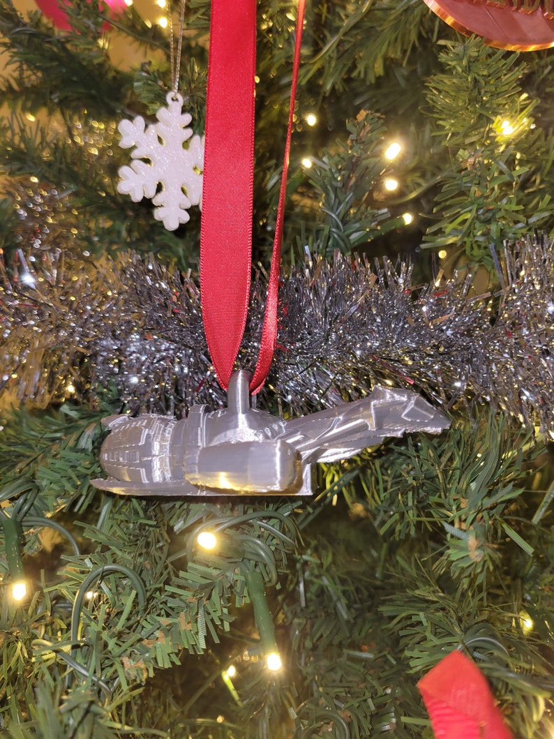 May include: A silver 3D printed ornament shaped like a spaceship hanging on a Christmas tree with a red ribbon and a white snowflake ornament.