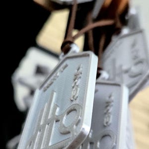 Puede incluir: Un conjunto de colgantes rectangulares plateados con caracteres japoneses y un logotipo, ensartados en cordones marrones. Los colgantes est&aacute;n enfocados, con otros ligeramente borrosos. El fondo es oscuro.