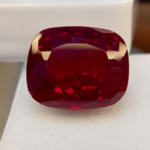 Può includere: Una gemma di rubino sfaccettata, taglio a cuscino. La gemma rosso intenso ha un alto grado di purezza, con sfaccettature che riflettono la luce, creando una brillantezza brillante. Il rubino è posto su uno sfondo bianco.