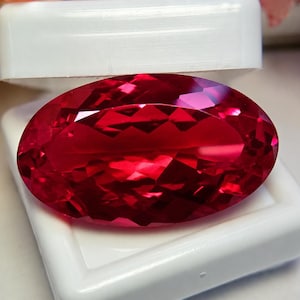 May include: A vibrant, oval-cut red gemstone with a faceted surface that reflects light, displayed resting inside a small, white, rectangular plastic storage box with the lid partially open, showcasing the deep crimson color and clarity.