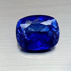 Peut inclure: Une pierre pr&eacute;cieuse taill&eacute;e en coussin, avec de multiples facettes, d'un bleu profond et vibrant. Les facettes r&eacute;fl&eacute;chissent la lumi&egrave;re, cr&eacute;ant un effet scintillant. La pierre est pos&eacute;e sur un fond gris clair neutre.