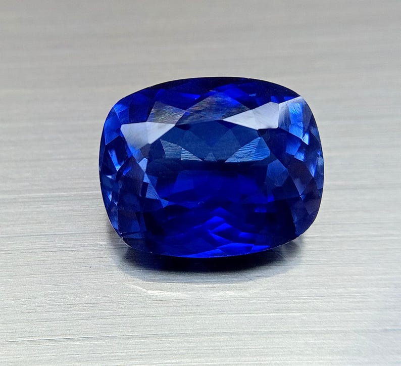 Peut inclure: Une pierre pr&eacute;cieuse taill&eacute;e en coussin, avec de multiples facettes, d'un bleu profond et vibrant. Les facettes r&eacute;fl&eacute;chissent la lumi&egrave;re, cr&eacute;ant un effet scintillant. La pierre est pos&eacute;e sur un fond gris clair neutre.