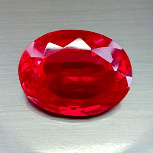 Può includere: Una gemma rossa vibrante, tagliata a forma ovale. Le sfaccettature della gemma catturano e riflettono la luce, mostrando la sua struttura interna. La superficie lucida della pietra presenta un colore rosso intenso e saturo.