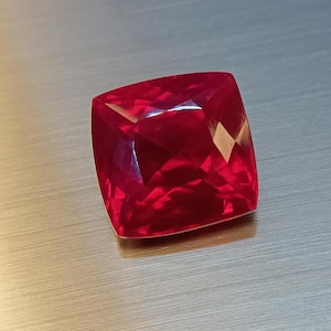 Könnte beinhalten: Ein facettierter, kissenförmiger Edelstein in tiefroter Farbe. Die Oberfläche des Edelsteins reflektiert das Licht und zeigt seine vielfältigen Facetten und seine innere Struktur. Der Edelstein ist vor einem neutralen Hintergrund platziert.