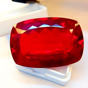 May include: A vibrant, cushion-cut red gemstone with a faceted surface that reflects light. The gemstone is positioned resting on an open, white plastic display box, showcasing its deep crimson color and geometric cut against the neutral container.
