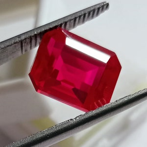 May include: A vibrant red gemstone with an emerald cut. The gemstone is held in a pair of tweezers.
